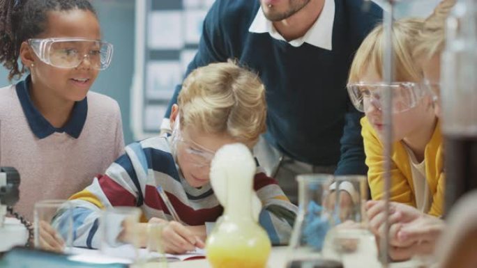 小学科学教室: 热情的老师向不同的孩子解释化学，小男孩在烧杯中混合化学物质，写下结果。孩子们有兴趣学