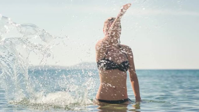 女人在海滩上享受夏天，站在海洋中，在炎热的晴天将海水溅向相机