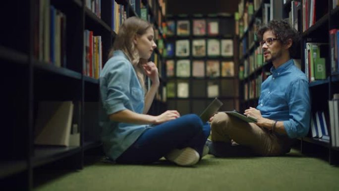 大学图书馆: 聪明的高加索女孩坐着，才华横溢的西班牙裔男孩盘腿坐在地板上，交谈，使用笔记本电脑并讨论