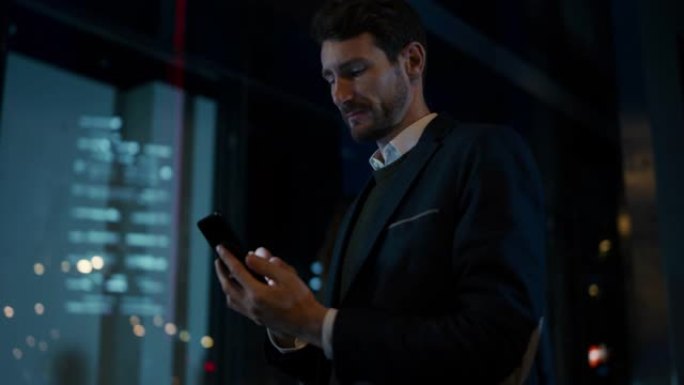 穿着西装的高加索商人晚上在黑暗的街道上使用智能手机。其他办公室人员走过。他看起来自信而成功。背景中的