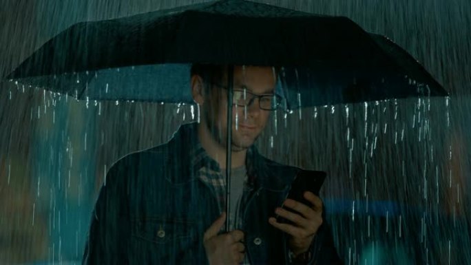 戴着眼镜的白人年轻人，穿着牛仔裤外套和方形衬衫，正在雨伞下使用智能手机。外面很黑，正在下雨。