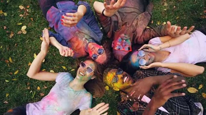 快乐的朋友的俯视图男性和女性躺在公园的草地上，脸上和衣服覆盖着明亮的油漆，看着相机和移动的手，微笑。