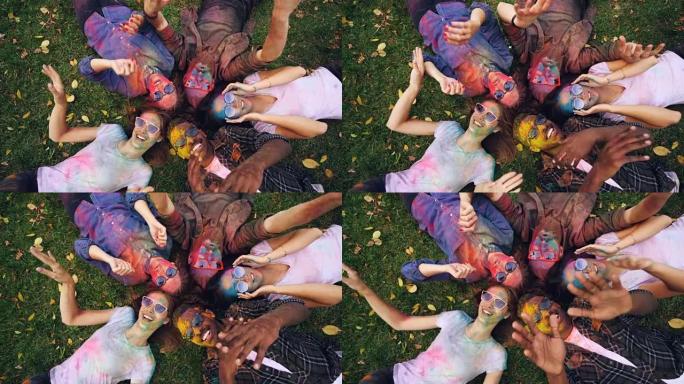 快乐的朋友的俯视图男性和女性躺在公园的草地上，脸上和衣服覆盖着明亮的油漆，看着相机和移动的手，微笑。