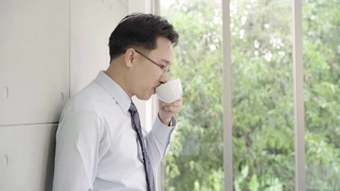 慢动作-成熟的亚洲商人喝着咖啡，从办公楼望向窗外的城市天际线。
