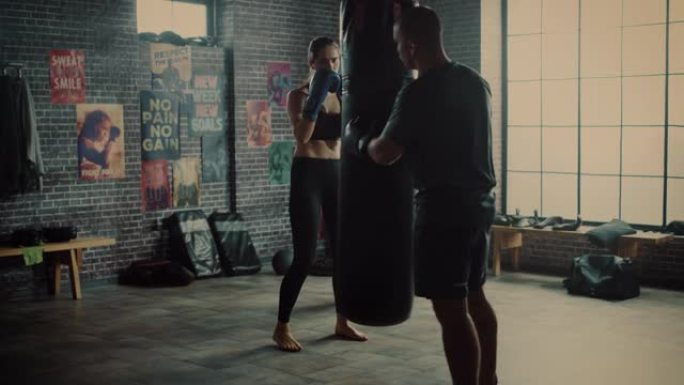 健身运动女性跆拳道运动员在阁楼健身房锻炼时拳打出气筒。她美丽而充满活力。强壮的教练拿着拳击包。激烈的