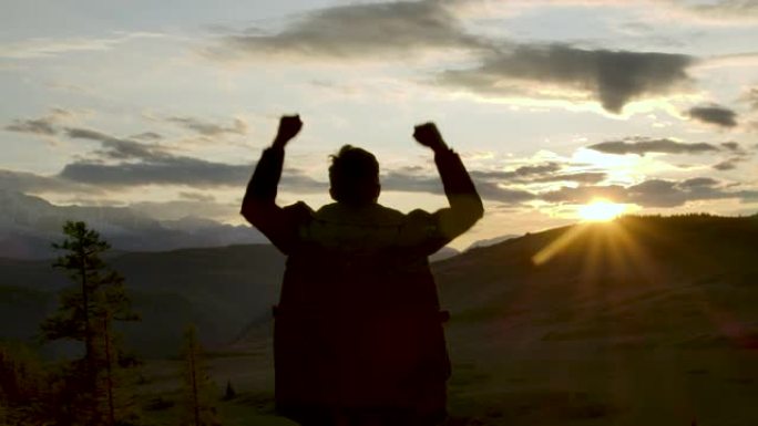 一个成功的快乐旅行男人站在欢乐之上，举起手来。快乐的剪影20多岁的男人看着山脉后面的日落天空。冒险旅