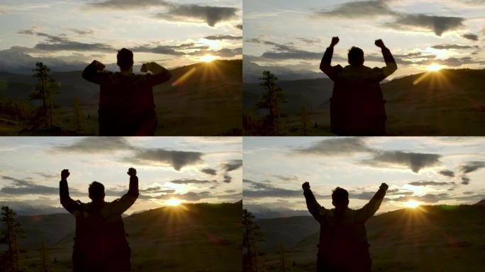 一个成功的快乐旅行男人站在欢乐之上，举起手来。快乐的剪影20多岁的男人看着山脉后面的日落天空。冒险旅