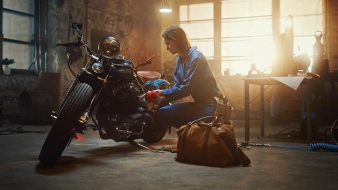 年轻漂亮的女机械师来到车库，开始在定制摩托车上工作。穿着蓝色连体裤的天才女孩。她用扳手拧紧螺母螺栓。
