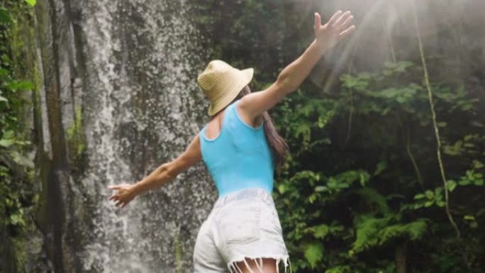 一个女人在一个瀑布前探索着世界上新的、神奇的、奇妙的地方，她被大自然包围着，伸开双臂呼吸和放松。