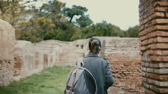 相机跟随年轻的女人背着背包在度假旅行中走在意大利Ostia Antica的古老红砖废墟中。