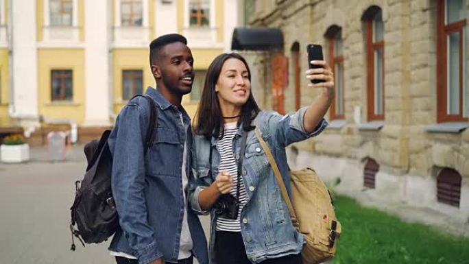 美丽的多种族夫妇正在与朋友在线聊天，使用智能手机站在街上背着背包进行视频通话。旅行和技术概念。