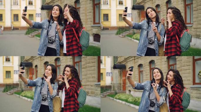 欢快的女孩外国旅行者正在使用智能手机站在户外进行自拍，并摆出手势，显示v字符号和手指的心脏并大笑。
