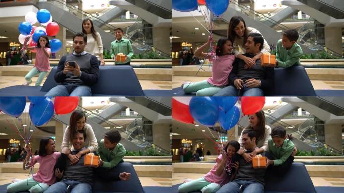 可爱的孩子和配偶令人惊讶的男人在购物中心用智能手机发短信，用气球和礼物