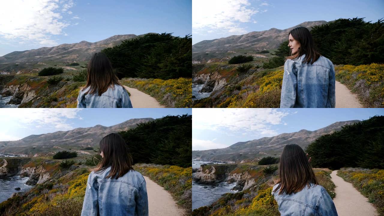 镜头跟随年轻快乐的女人在黄色的小花路上走向大苏尔海岸线的岩石和森林。
