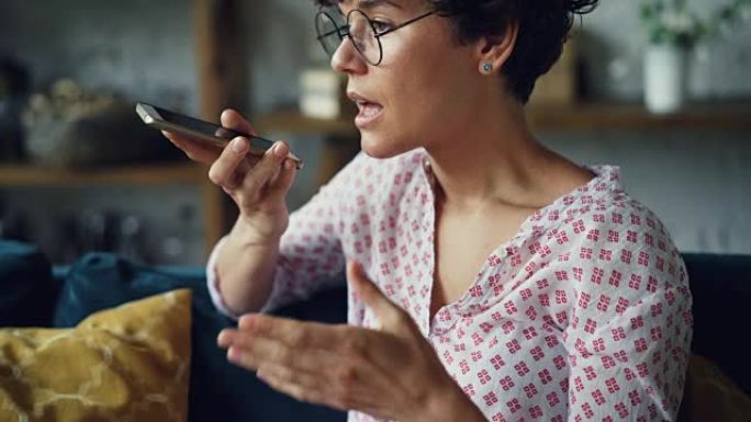 戴着眼镜和休闲服的漂亮黑发女人正在触摸智能手机屏幕，然后手持会说话的手持设备。扬声器模式或录音机概念