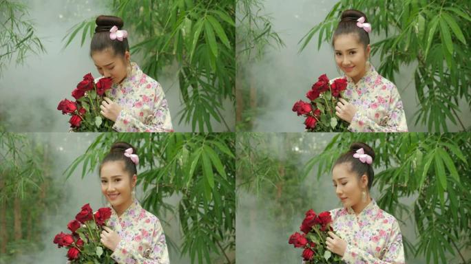 特写传统服装迷人的亚洲女性的肖像，一束红玫瑰的鲜花看着镜头，在森林的背景上微笑。