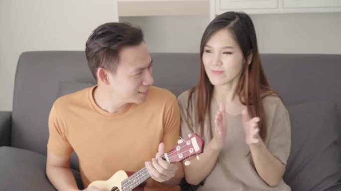 亚洲夫妇在家里的客厅弹吉他和唱歌，甜蜜的夫妇在家里放松时躺在沙发上享受爱情时刻。生活方式夫妇在家放松