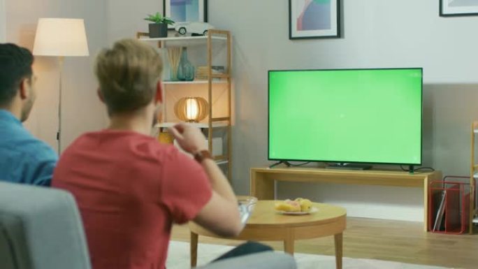 在客厅里，两个朋友坐在沙发上，拿着控制器，在绿色色度键电视屏幕上玩竞争性视频游戏。一个朋友预告另一个