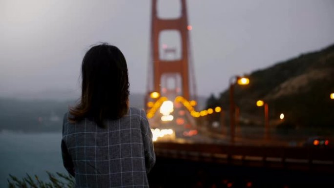 后视图年轻的千禧一代学生女孩独自站在黑暗的夏夜观看标志性的金门大桥交通