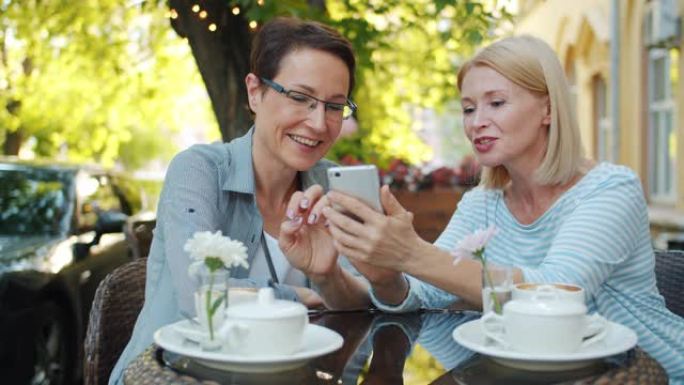 快乐的女士们笑着在户外咖啡馆看着智能手机屏幕聊天