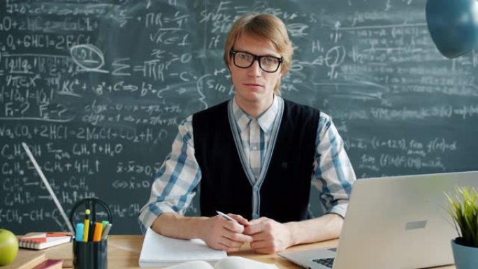 戴着眼镜的严肃年轻人的肖像看着坐在教室桌子上的相机