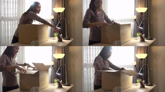 漂亮的年轻亚洲女人在家站在明亮的客厅的桌子上打开纸箱，在线送货
