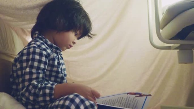 亚洲男孩在毯子堡垒下用手电筒阅读童话
