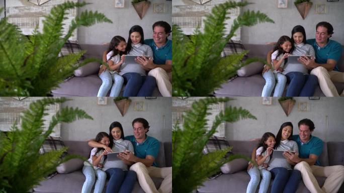 美丽的怀孕母亲，丈夫和女儿坐在沙发上看着平板电脑上的视频微笑