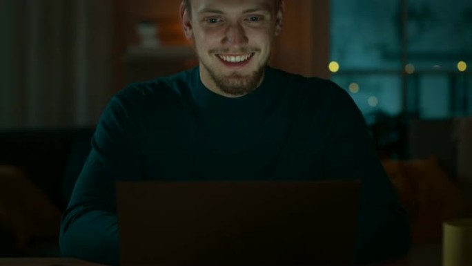 英俊的微笑男人坐在家里的办公桌前在笔记本电脑上工作。一位年轻的自由职业者的肖像在他舒适的客厅里用电脑