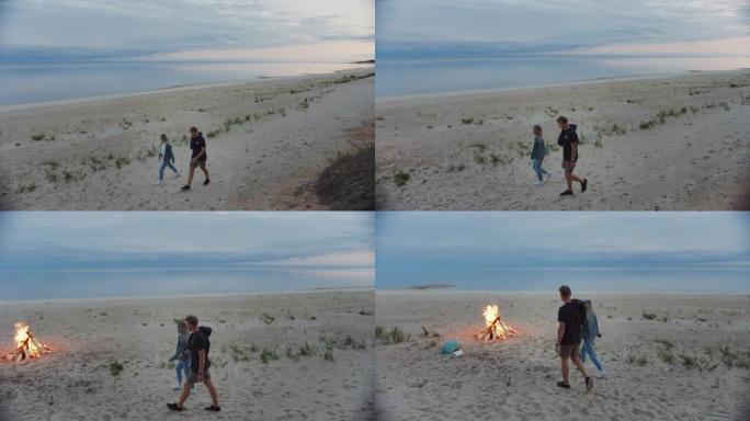 幸福的年轻夫妇走在荒芜的海滩上。黄昏时分平静的大海。他们走向篝火。女孩拿着啤酒瓶，年轻人穿着短裤。夏