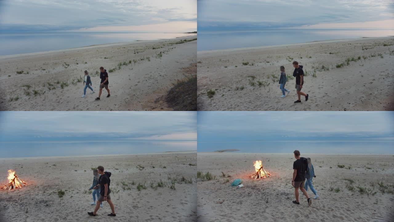幸福的年轻夫妇走在荒芜的海滩上。黄昏时分平静的大海。他们走向篝火。女孩拿着啤酒瓶，年轻人穿着短裤。夏