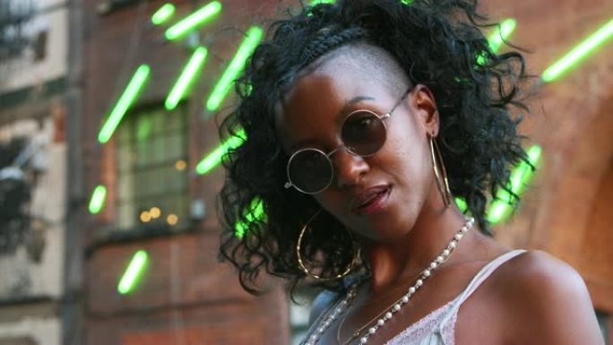 时尚的年轻黑人女性戴着圆形太阳镜，在城市街道上对着相机摆姿势，特写，低角度，背景中模糊的霓虹灯