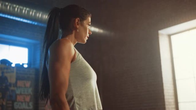 一个美丽强壮的黑发女人站在阁楼工业健身房的肖像，上面贴着激励海报。经过激烈的健身训练后，她喘不过气来