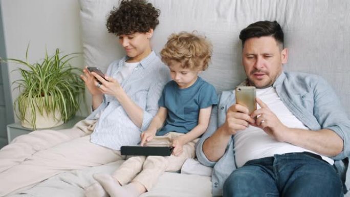 孩子在家床上在平板电脑上玩游戏时，男人和女人使用智能手机