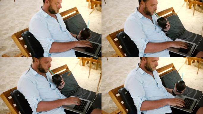 认真专注的年轻商人坐在海边的休闲沙滩椅上，用笔记本电脑喝椰子水果鸡尾酒。