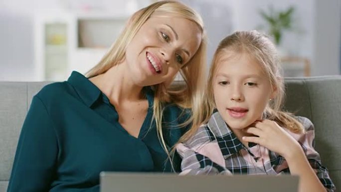 美丽的年轻妈妈和她可爱的小女儿坐在家里的沙发上使用笔记本电脑。一家人花时间在电脑上一起看视频和卡通。