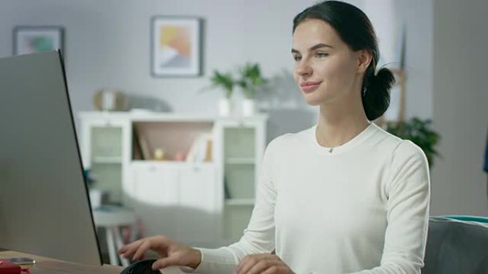 美丽的年轻女子在舒适的客厅在个人电脑上工作的肖像照片。她迷人地微笑着。