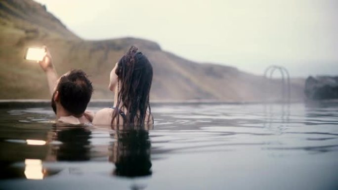 年轻夫妇在山谷的智能手机上拍照。在冰岛的温泉里游泳的男人和女人。