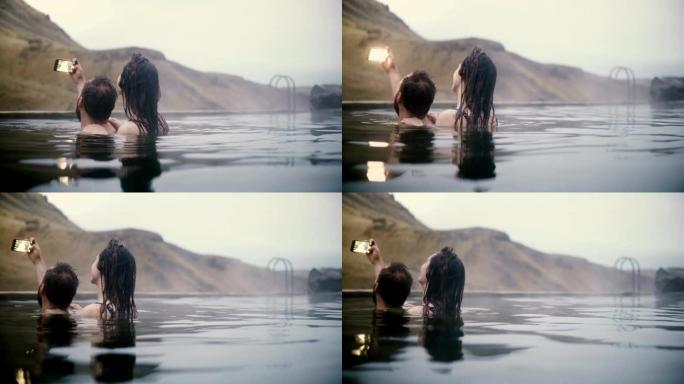 年轻夫妇在山谷的智能手机上拍照。在冰岛的温泉里游泳的男人和女人。