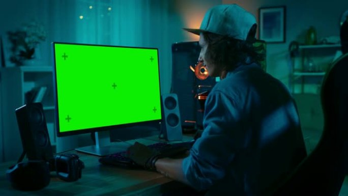 专业玩家在他功能强大的个人电脑上玩在线视频游戏，配有彩色霓虹灯发光二极管灯。绿屏模型。年轻人戴着帽子