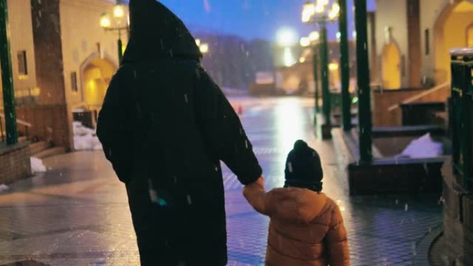 孩子在城市街道上与母亲一起在夜晚与雪同行。