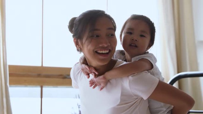 微笑的亚洲妈妈背着孩子的女儿在床上嬉笑