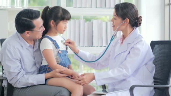 女儿科医生用听诊器检查可爱的小女孩。孩子开心地微笑着指着医生。父亲抱着她的孩子。中国和香港的医疗系统