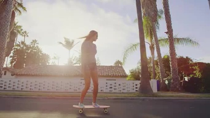 迷人的冒险女孩在日落时沿着棕榈树成荫的街道滑板