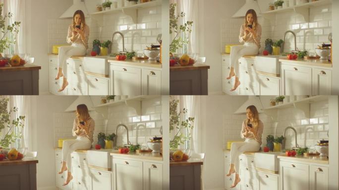 穿着条纹套头衫和白色裤子的美丽年轻女性正坐在厨房家具上，用她的智能手机在一个现代阳光明媚的厨房里。她