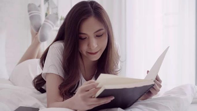 美丽迷人的亚洲女人躺在床上看书的肖像，在家里的卧室里放松。生活方式女性使用在家放松时间的概念。