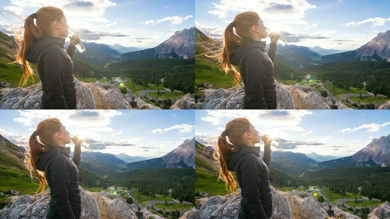 女人在日落时俯瞰山谷的山顶上从水瓶中喝水