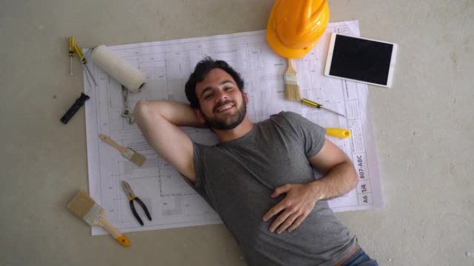 英俊的建造者躺在蓝图上的地板上，旁边放着工具和平板电脑，对着镜头微笑