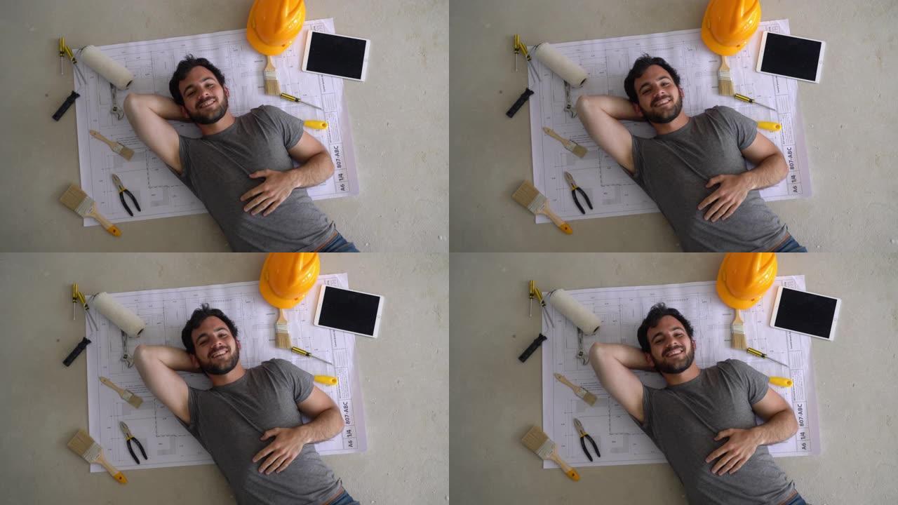 英俊的建造者躺在蓝图上的地板上，旁边放着工具和平板电脑，对着镜头微笑