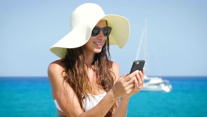 一个美丽的年轻女子微笑着，在电话里写了一条信息，白色比基尼，太阳镜，帽子，大海的背景。